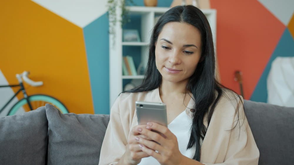Una mujer revisa opciones en su teléfono, sentada en el sofá de su casa.